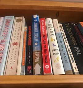 A close up of a bookshelf with a dozen books of all genres--like Gloria Steinem's My Life on the Road, Suketu Mehta's This Land is Our Land, Rolling Stone's biography of Johnny Cash, and more.